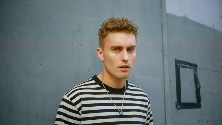 Sam Fender looks at the camera, wearing a black and white striped tee and a chain with a ring on it