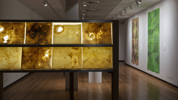 A collection of artworks is displayed on a lightbox, a green and a yellow modern art painting line the white wall facing the lightbox