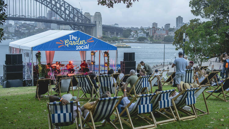 Rows of stripy deck chairs on a grassy lawn in front of a stage overlooking the Sydney Harbour Bridge.