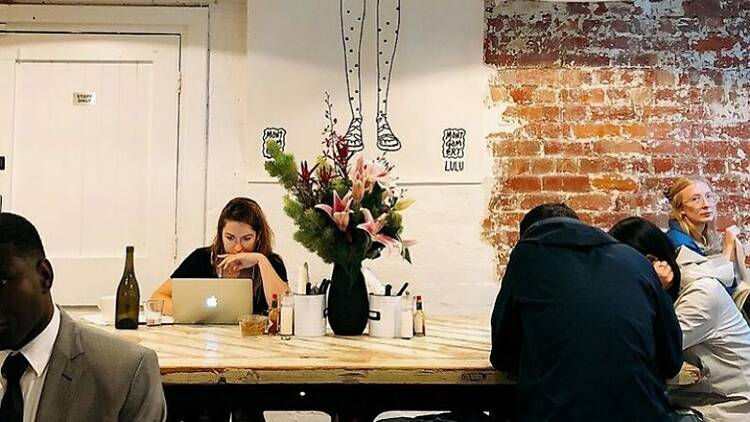 Several people sit around a common table at a cafe, one woman is on a latptop. There is an exposed brick wall and a mural of a girl in high socks.
