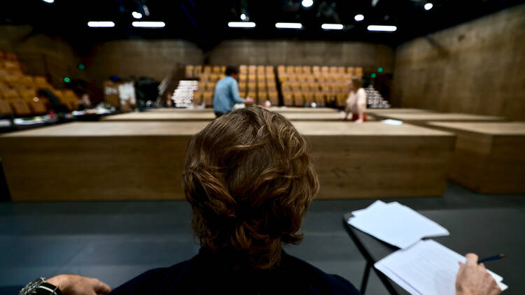 A close-up of the back of a man's head, with people working on an unfinished theatre in the background.