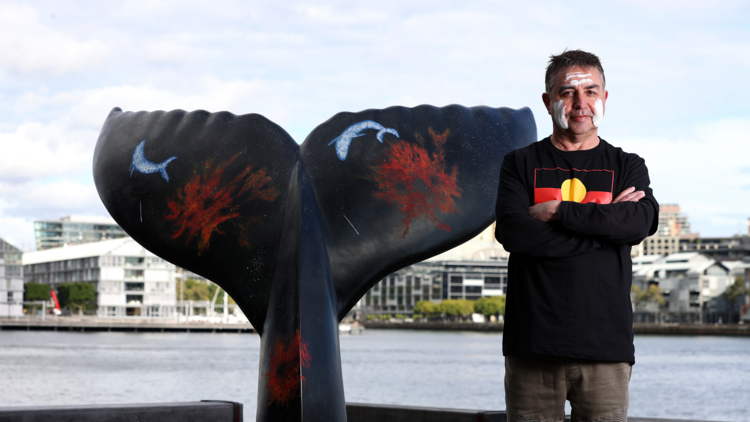 A man with white paint on his face and wearing a black t-shirt with the Aboriginal flag on it stands with his arms crossed next to a black sculpture of a whale's tail.