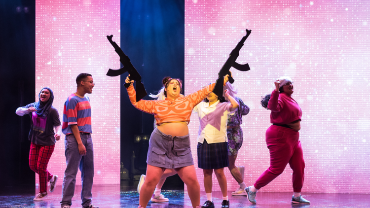 A woman on stage throws her arms in the air holding 2 fake rifle cutouts 