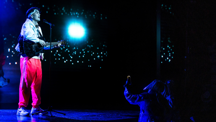 An actor sings into a microphone on stage dressed as a boyband lead singer against a backdrop of glittering lights