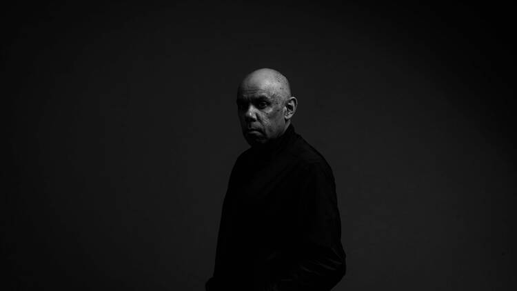 A black and white image of a bald man in a black skivvy against a dark backdrop.