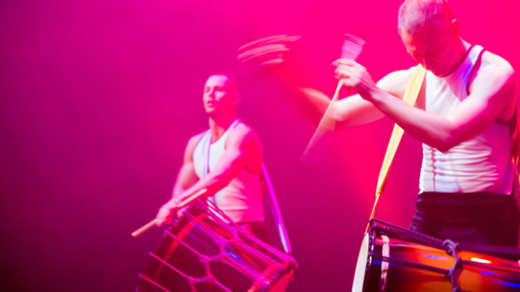 Two men in white singlets play drums strapped to their bodies under a pink-tinted light.