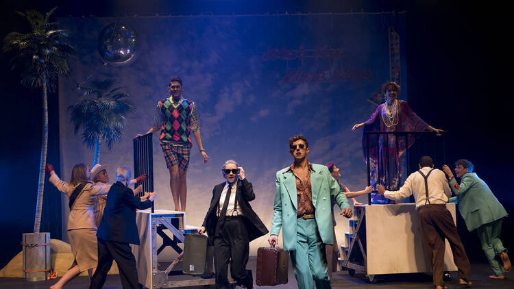 A cast of actors pose on stage in front of a cloud backdrop and prop palm trees.