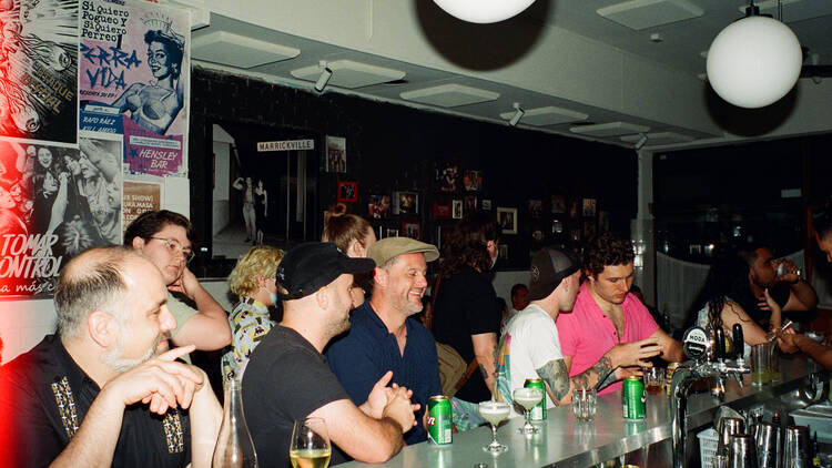 A crowd of people standing and sitting around a long bar, with posters on the wall in the background.