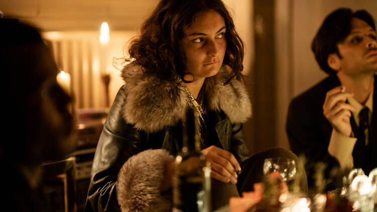 A woman sits at a dinner table in a jacket lined with fur. She bites her lip, and leans towards the table