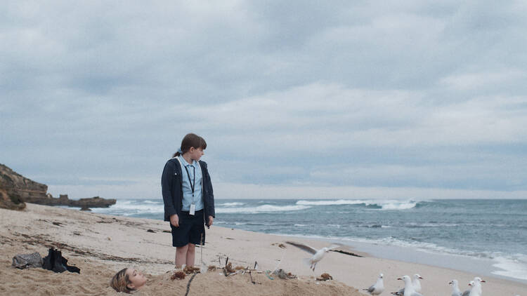 A still from Moja Vesna: a young girl stands on the beach next to her sister, buried in sand up to her head, it's a stormy sky.