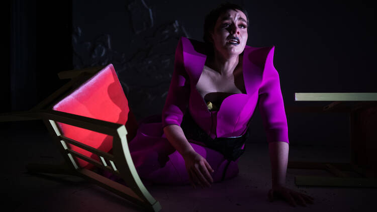 A woman sits on the floor next to a toppled chair. She is wearing an angular purple suit and looks anguished