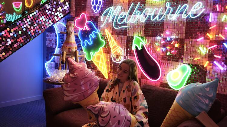 A woman sitting on a couch with ice cream balloons in front of a wall of neon signs.