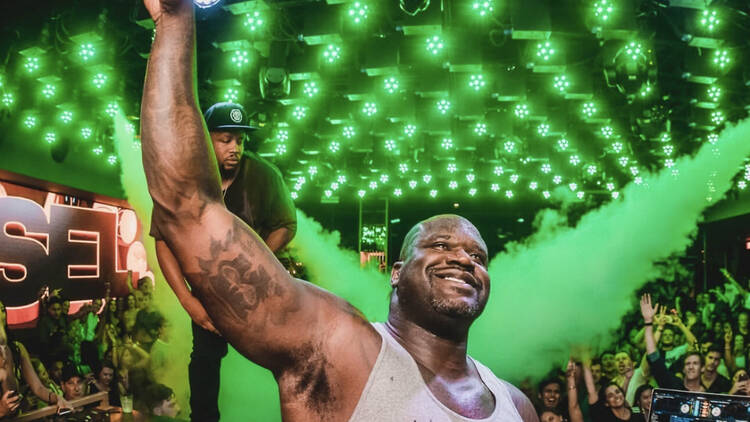 Shaquille O'Neal, as DJ Diesel, holds his hand in a peace sign, as a crowd dances behind him in green laser lights