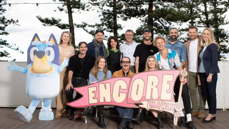 A large group of people pose for a photo with a life-size cardboard cut-out of Bluey and a sign that says 'Encore Manly'.