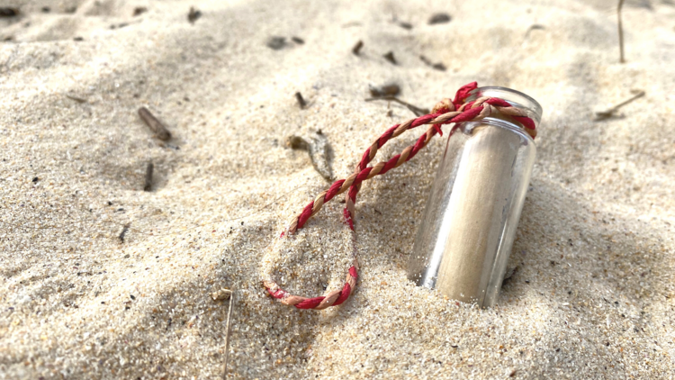 A tiny glass vial with a rolled up piece of paper in it lies half buried in sand