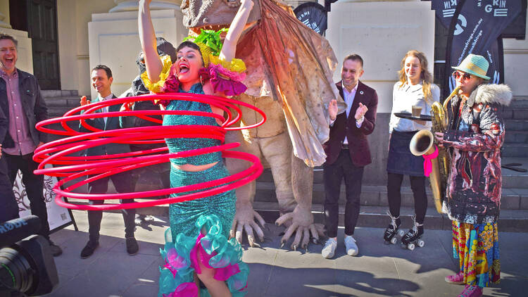 A woman in a colourful outfit is spinning multiple hula hoops as people playing instruments stand behind her