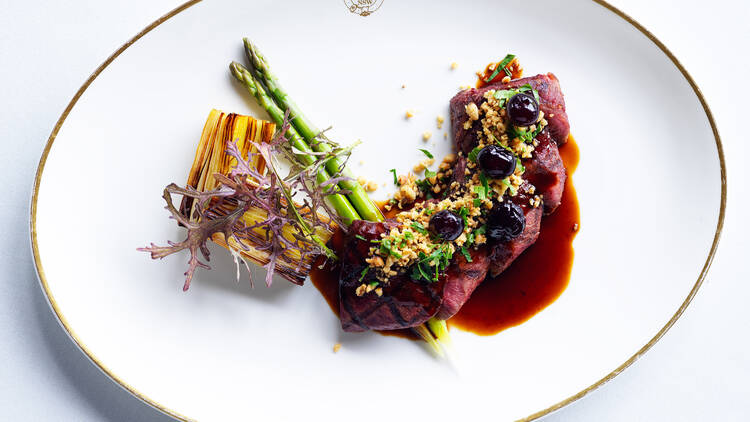 An expertly plated meal of meat, vegetables and sauce sitting on a white oval serving dish with gold trim.