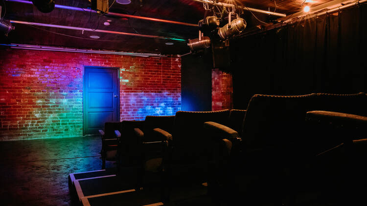 The interior of a performance space features large lounge-like chairs looking onto a multi-coloured lit stage area