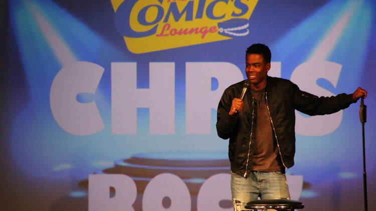 Chris Rock stands on stage, backed by a huge banner with his name and The Comics Lounge logo