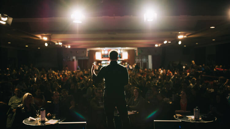 A comedian stands on stage, gesturing to a crowd. We see their back, and look out into the packed audience.
