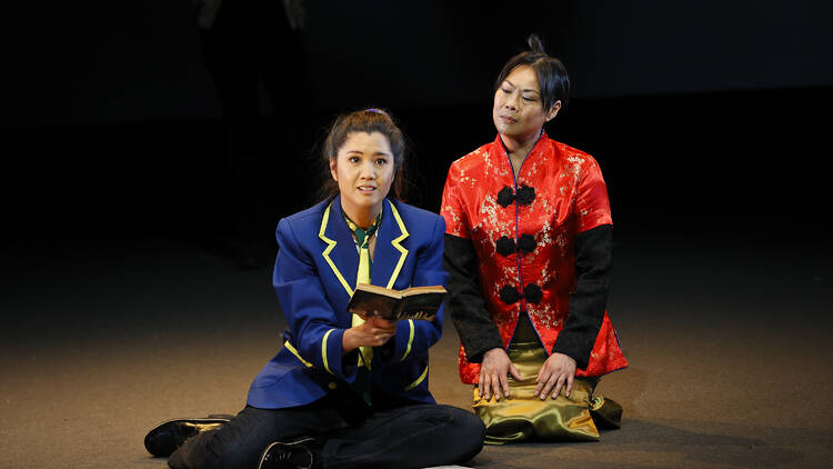 A girl in a school uniform sits on the ground, holding an open book, as a woman in traditional Vietnamese dress looks on.