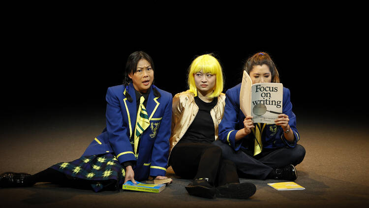 Three girls sit on the floor. One in school uniform speaks toward the audience and the other reads a book. A third, in a gold jacket and black outfit, looks annoyed.