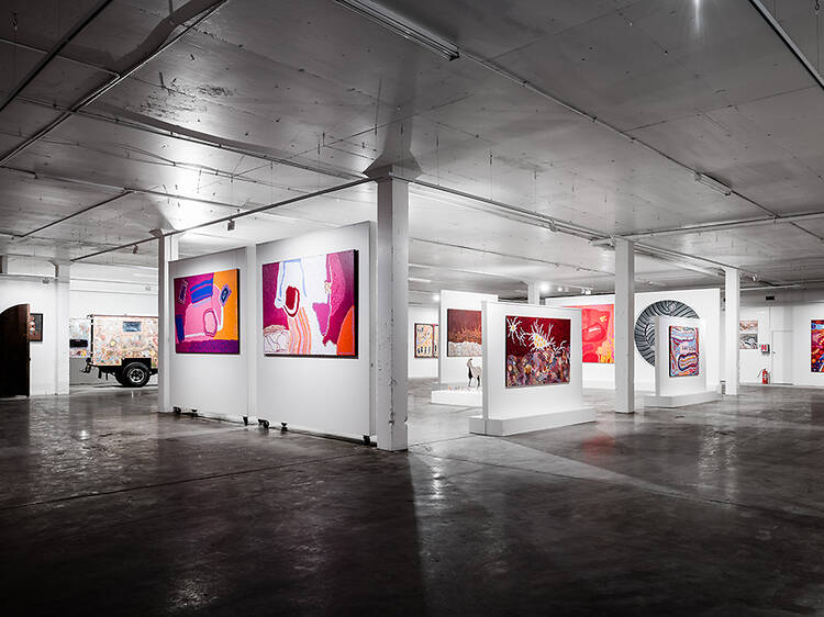 A gallery space houses many colourful artworks on white partition walls in pinks and reds