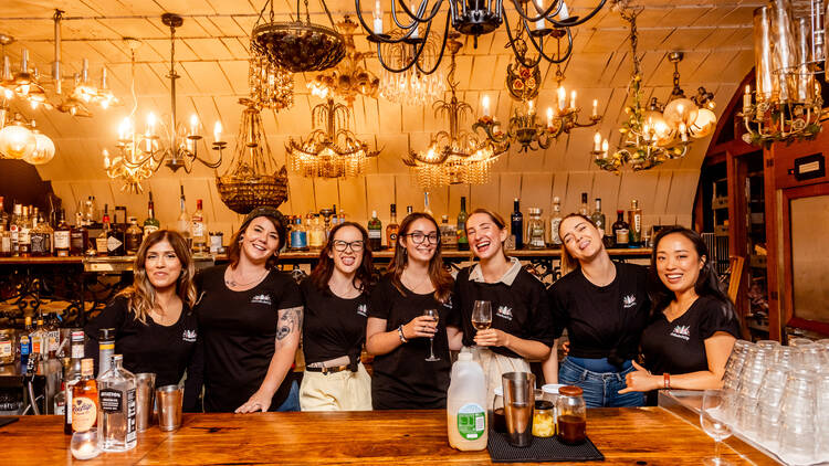A group of people all wearing matching black t-shirts stand behind a bar lined with alcohol. 