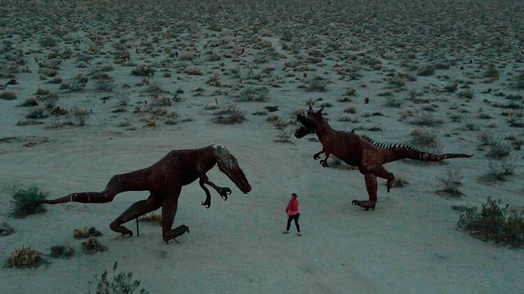 A woman walking through the desert, between two dinosaur sculptures.