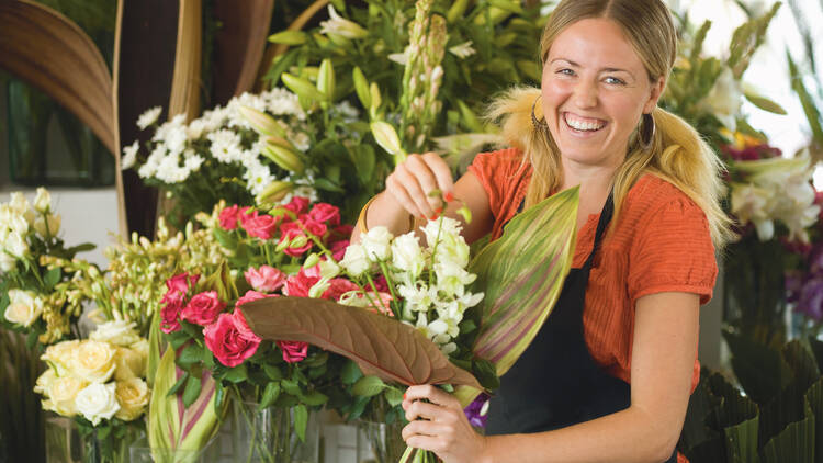 A smiling blonde woman with pigtails and wearing a black apron holds a big bunch of flowers.