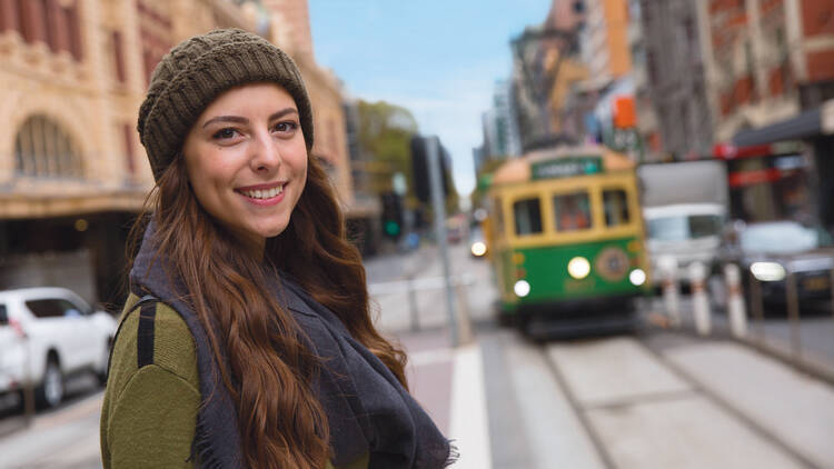 A smiling brunette girl wearing a beanie stands at a tram stop as an old-school Melbourne tram approaches.
