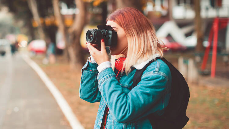 A girl with ombré hair and wearing a denim jacket holds a camera up to her face to take a picture.