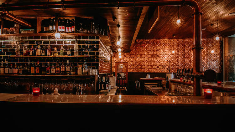 An industrial bar is lined with bottles of alcohol, and features decorative tiles on the wall in the distance