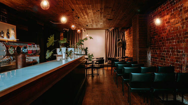 A brick lined bar features dark chairs and tables, a wooden bar with a glass top, and red-tinged lighting