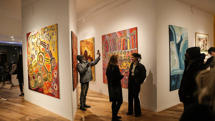 A gallery with white walls features indigenous artworks and pottery, people are wandering through