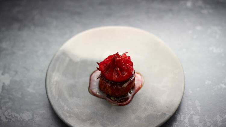 A grey plate sits on a textured grey table, it's adorned with a brilliant red circular lump of pate, topped with a red flower