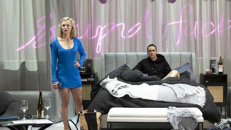 A set is constructed like a bedroom. A woman in a short blue dress stands next to the bed, a man in a black hoodie sits on the bed, looking into the audience