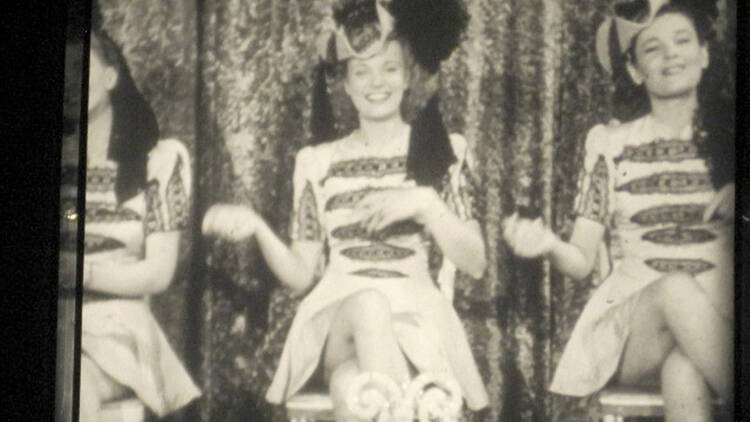 A sneak peek at one of the vintage 16mm films you'll see at the event. Three women sit in chairs and appear to lift they're arms as if they're dancing.