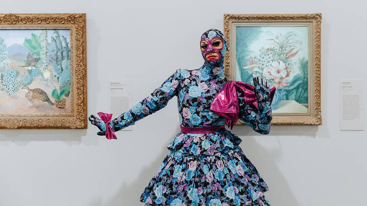 A person in a floral blue-toned ball gown stands in front of paintings in a gallery. The ball gown extends into a balaclava and gloves.