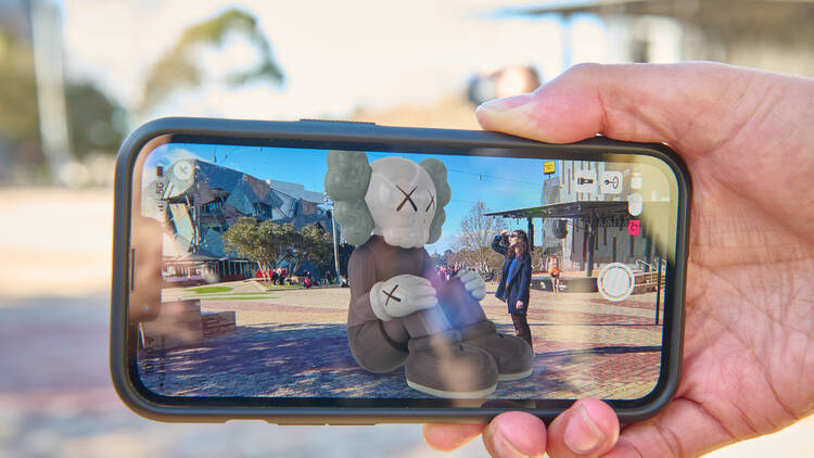 A person holds up a phone with the camera opened. A virtual Kaws sculpture appears next to the person in the photo