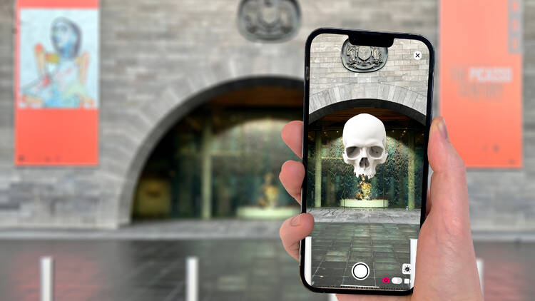 A person holds up a camera on a phone, and a giant virtual skull appears above the entrance to the NGV