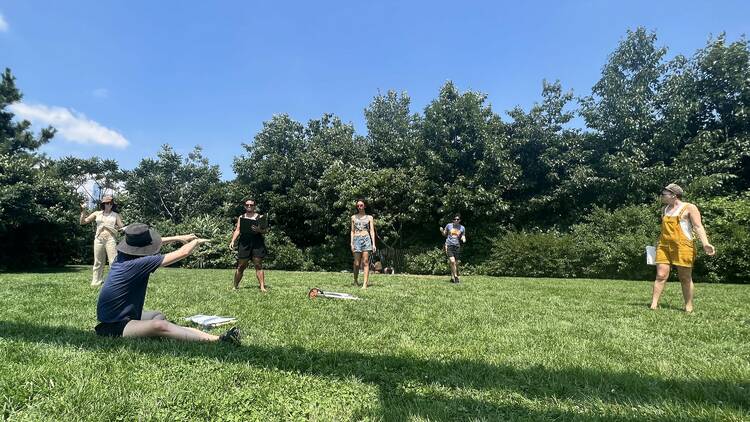 Six actors practice the play on a grassy patch.