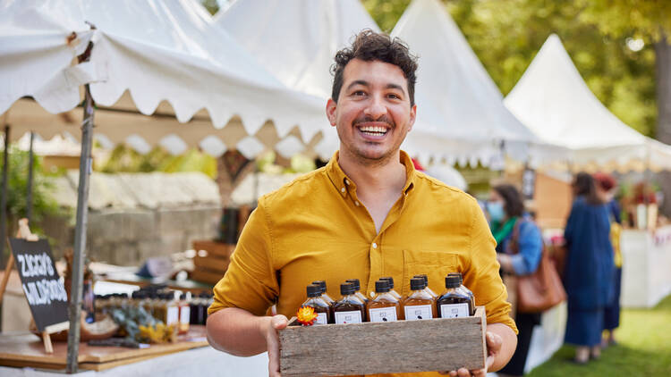 A smiling man in a yellow shirt holds a wooden box of juices at a market.