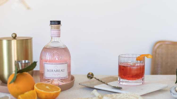 A pink gin bottle on a table next to a Negroni and half-cute oranges.
