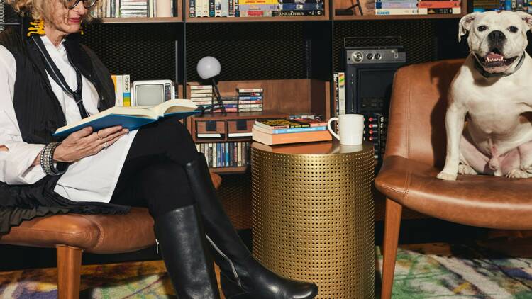 A woman sits in a chair reading a book. A dog sits in the chair next to her.