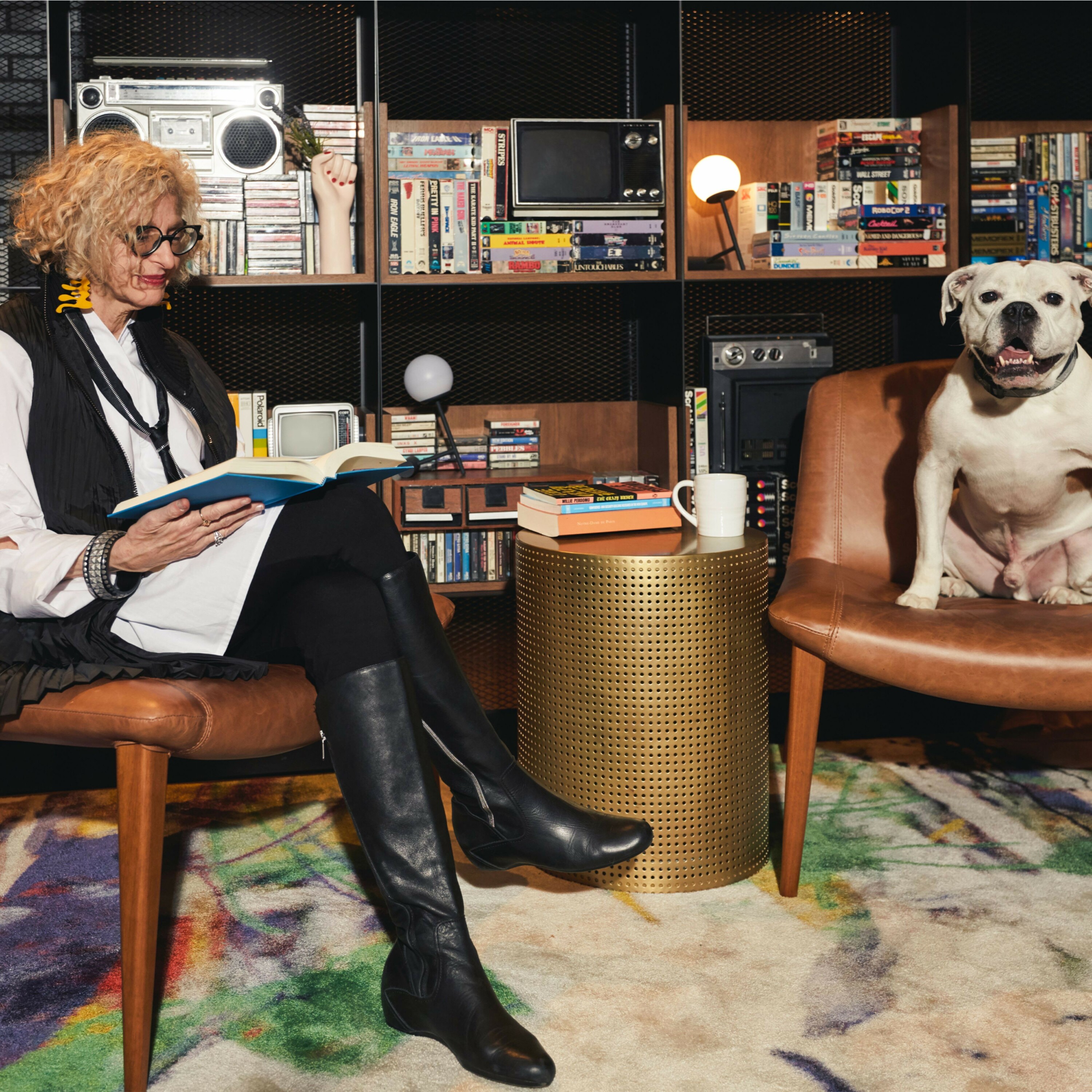 A woman sits in a chair reading a book. A dog sits in the chair next to her.