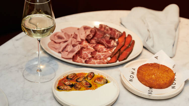 A white marble table features a salumi plate, glass of white wine, a fried circular patty on a plate emblazoned with Nomad