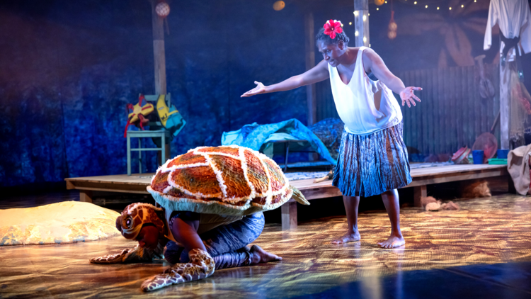 A performer holds her hands up in exasperation as a performer in a turtle costume crawls across the stage