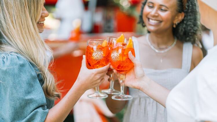 A group of three girls are smiling and drinking an Aperol Spritz