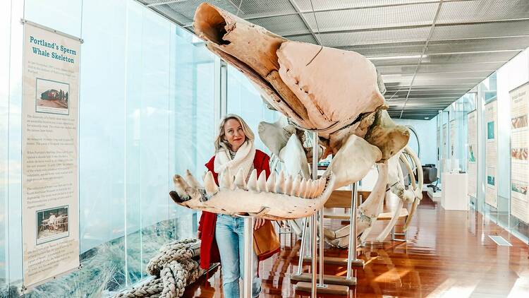 A blonde woman in a red coat stands next to a large sperm whale skeleton in a museum.